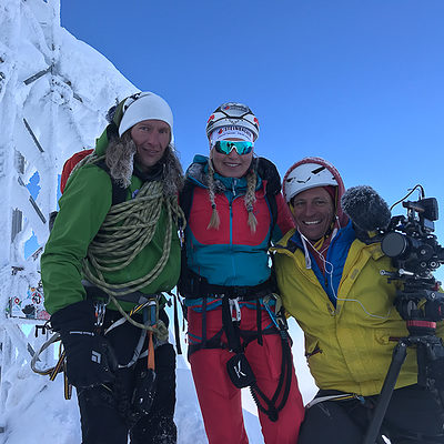 Drei Bergsteiger am verschneiten Gipfelkreuz, professionelle Kamera für Filmproduktion dabei