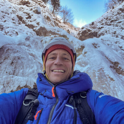 Mann mit Helm und blauer Jacke macht Selfie beim Winterwandern in verschneiter Berglandschaft