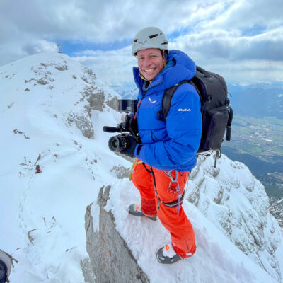 Kameramann mit Helm filmt auf schneebedecktem Berggipfel, professionelle Luftaufnahmen